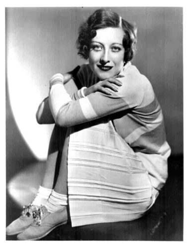 Black-and-white portrait of a woman with short wavy hair, wearing a striped dress, sitting with arms around her knees and smiling.