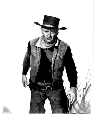 Black-and-white photo of a rugged cowboy wearing a wide-brimmed hat, vest, scarf, and belt, standing in a desert setting and looking ahead.