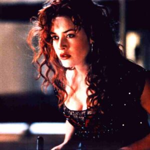Woman with curly red hair in a sparkling black top leans forward, looking intently to the side in a dim, moody setting.