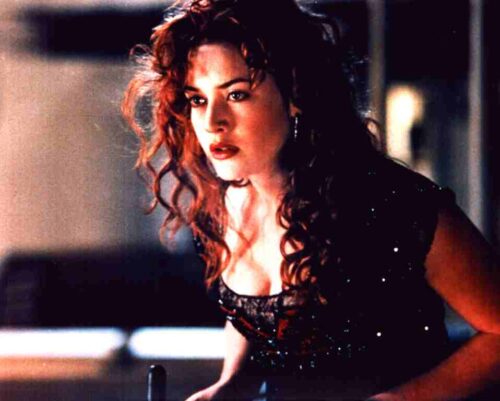 Woman with curly red hair in a sparkling black top leans forward, looking intently to the side in a dim, moody setting.