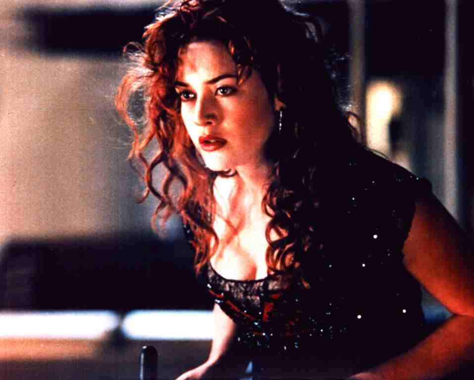 Woman with curly red hair in a sparkling black top leans forward, looking intently to the side in a dim, moody setting.