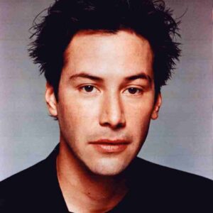 Head-and-shoulders portrait of a man with spiky dark hair, wearing a black shirt, against a neutral gray background.