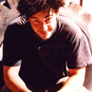Young man with dark, messy hair wearing a black t-shirt, leaning toward the camera with a friendly smile.