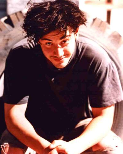 Young man with dark, messy hair wearing a black t-shirt, leaning toward the camera with a friendly smile.