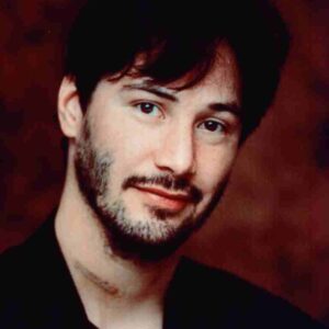 Portrait of a man with dark hair and light stubble, wearing black, looking at the camera against a brown background.