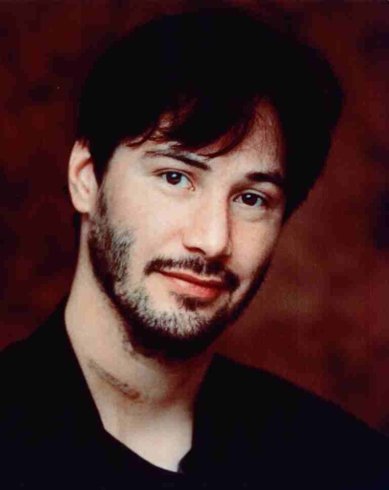 Portrait of a man with dark hair and light stubble, wearing black, looking at the camera against a brown background.