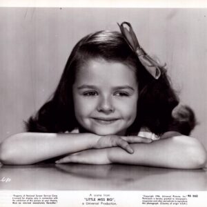 Young girl with a bow in her hair rests her chin on folded arms and smiles at the camera in a black-and-white portrait.