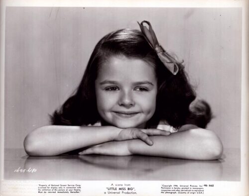 Young girl with a bow in her hair rests her chin on folded arms and smiles at the camera in a black-and-white portrait.