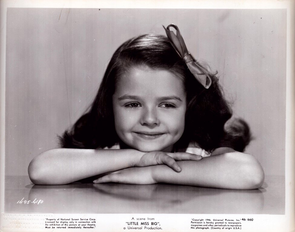 Young girl with a bow in her hair rests her chin on folded arms and smiles at the camera in a black-and-white portrait.