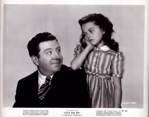 A man in a suit smiles at a young girl in a striped dress who rests her head on her hand and looks away.