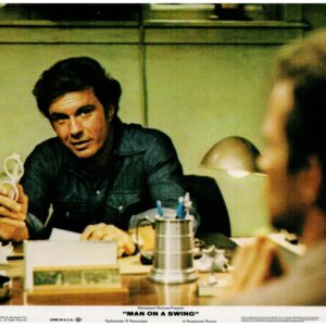 Actor in a denim shirt sits at a cluttered desk, extending a small looped wire toward someone off-camera in a dim, office-like setting.