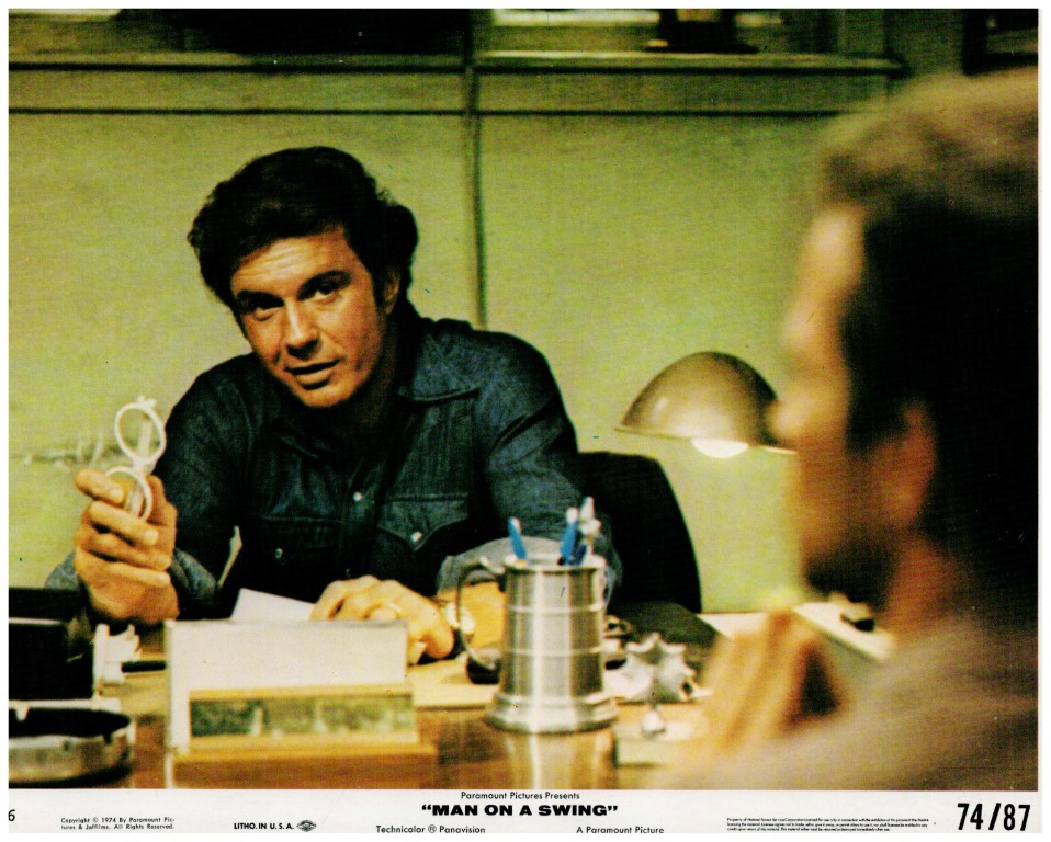 Actor in a denim shirt sits at a cluttered desk, extending a small looped wire toward someone off-camera in a dim, office-like setting.