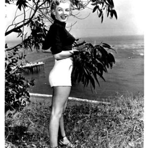 Vintage-style photo of a smiling woman in a black top and white shorts posing beside coastal vegetation by the sea.