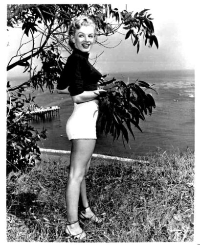 Vintage-style photo of a smiling woman in a black top and white shorts posing beside coastal vegetation by the sea.