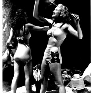 Two women in vintage swimsuits on a sunny beach, one swinging a rope while the other faces away, with beach gear around them.