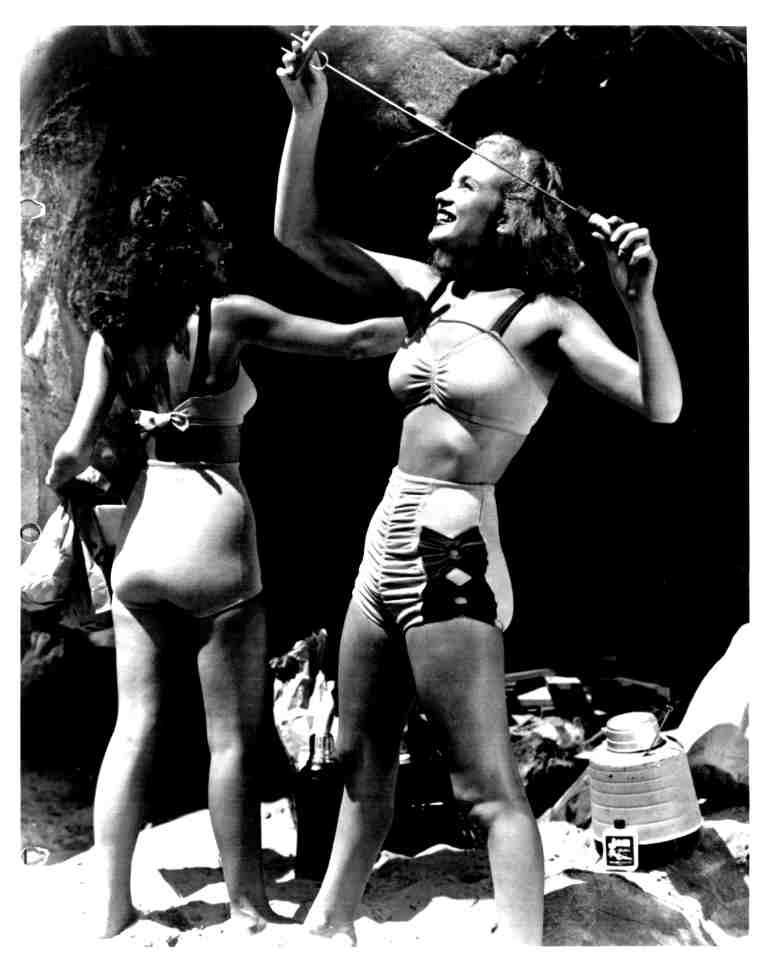 Two women in vintage swimsuits on a sunny beach, one swinging a rope while the other faces away, with beach gear around them.