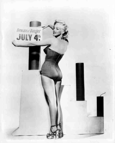 Pinup model in a one-piece swimsuit posing beside a sign that reads 'JULY 4th' and 'Beware the danger'.