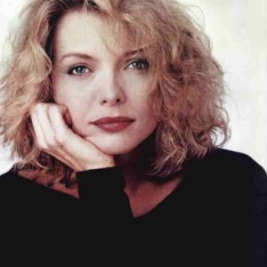 Portrait of a woman with shoulder-length curly blonde hair, resting her chin on her hand, wearing a black top against a light background.