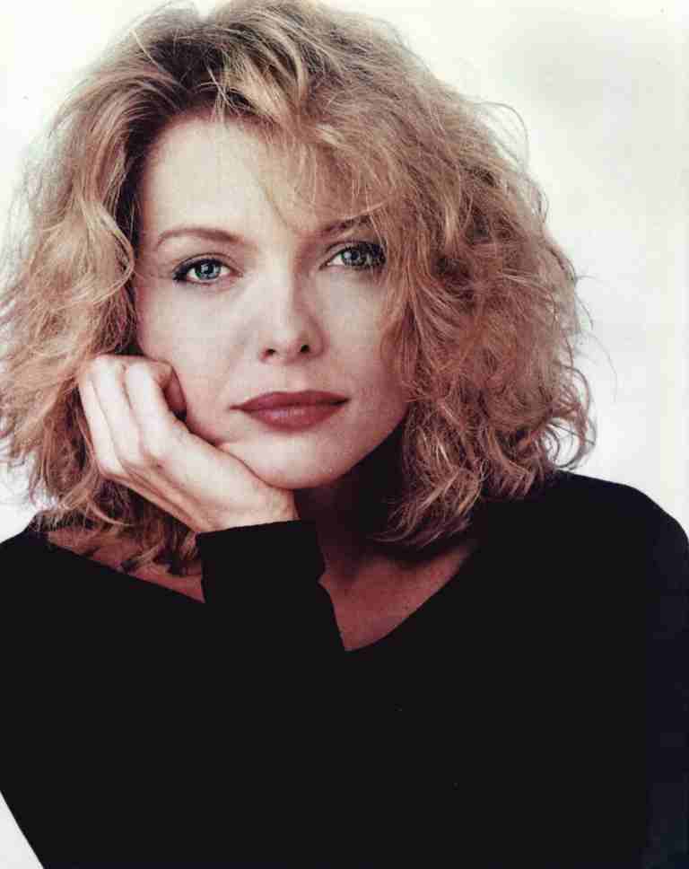 Portrait of a woman with shoulder-length curly blonde hair, resting her chin on her hand, wearing a black top against a light background.