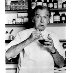 Older man in a lab coat stirs a dark liquid in a glass bowl, with bottles on shelves behind him in a cluttered lab.