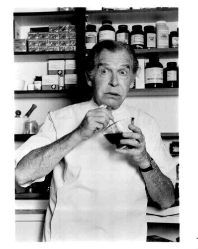 Older man in a lab coat stirs a dark liquid in a glass bowl, with bottles on shelves behind him in a cluttered lab.