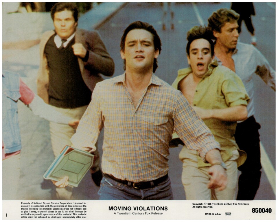 Promo still from the 1985 film Moving Violations: three men run toward the camera on a street, central figure in a plaid shirt carrying a lunchbox, movie title area at bottom.