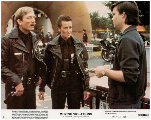 Two police officers in black leather jackets talk with a man who’s taking notes beside a parked car at an outdoor event.