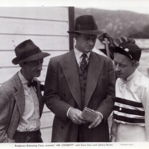 Promotional still for the film Mr. Celebrity, showing three men in fedoras and suits gathered around a newspaper.