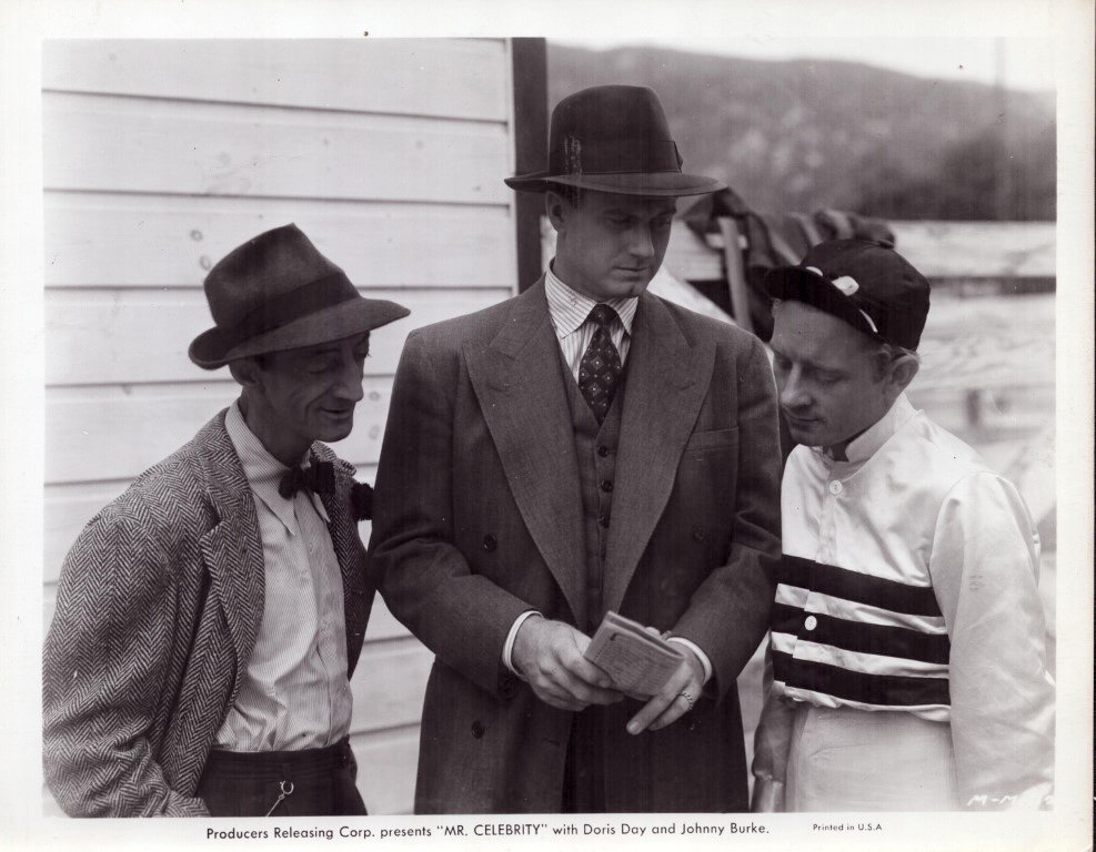 Promotional still for the film Mr. Celebrity, showing three men in fedoras and suits gathered around a newspaper.