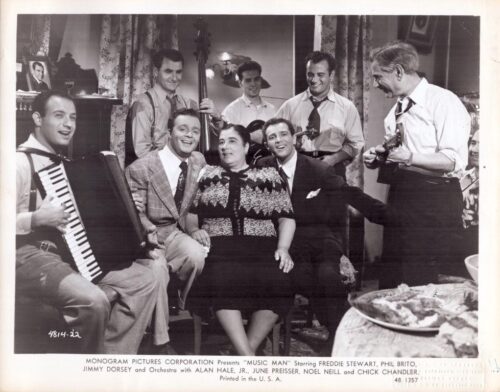 Black-and-white photo of a mid-20th-century living-room band: accordion player, bass, and guitarists surround a smiling couple seated in the center.