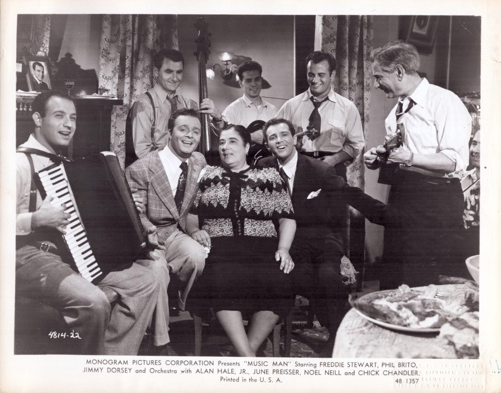 Black-and-white photo of a mid-20th-century living-room band: accordion player, bass, and guitarists surround a smiling couple seated in the center.