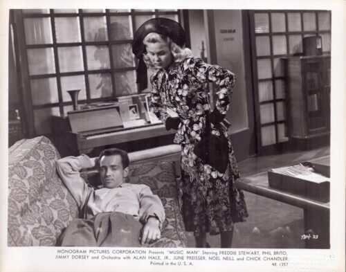 Promotional still from the 1950s film Music Man: a man lounging on a patterned sofa while a woman in a floral dress and wide hat leans over him in a living room set.