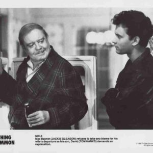 Black-and-white still of two men by a car: an older man in a plaid robe holds a cigarette and smiles at a younger man in a dark jacket.