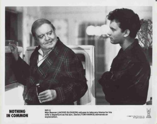 Black-and-white still of two men by a car: an older man in a plaid robe holds a cigarette and smiles at a younger man in a dark jacket.