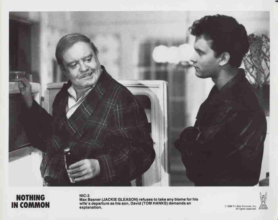 Black-and-white still of two men by a car: an older man in a plaid robe holds a cigarette and smiles at a younger man in a dark jacket.