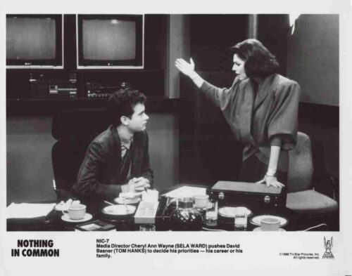 A woman stands beside a seated man at a cluttered desk with coffee cups, gesturing as they talk in a black-and-white office scene with monitors behind them.