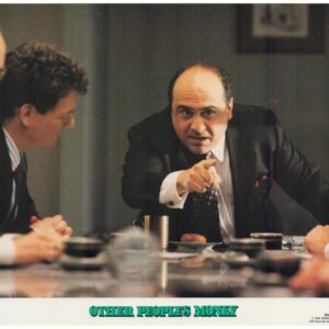 Businessman in a suit leans forward, pointing at someone across a conference table during a tense meeting.