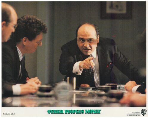 Businessman in a suit leans forward, pointing at someone across a conference table during a tense meeting.