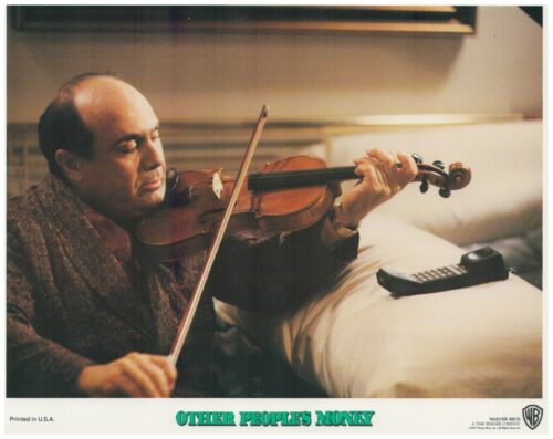 Man seated on a couch plays a violin with a bow; a remote rests on a white pillow nearby, on a lobby card for 'Other People's Money'.
