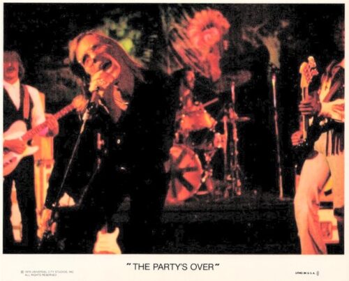 Live rock band performing on stage with lead singer center and red-tinted lighting; drummer in back. Caption reads 'The Party's Over' (album promo).