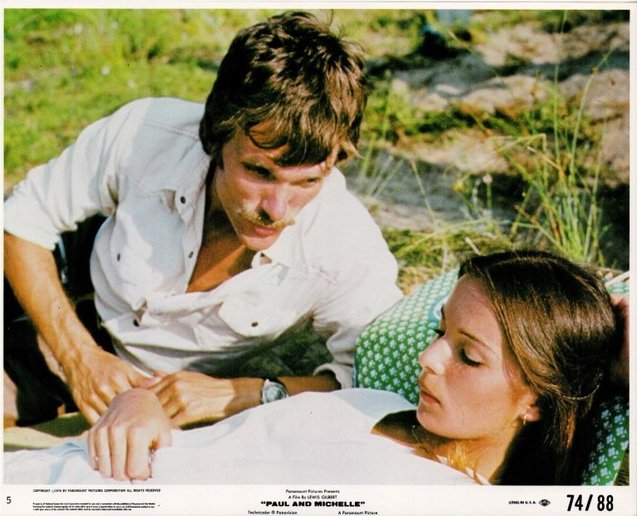 Man with a mustache leaning over a reclining woman outdoors, both looking at each other.