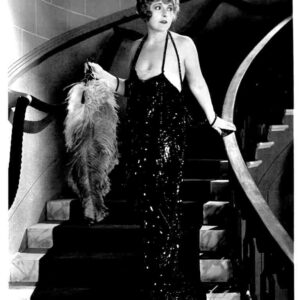 Black-and-white photo of a glamorous woman in a glittering halter-neck evening gown standing on a curved staircase, holding a feathered boa and looking off to the side.