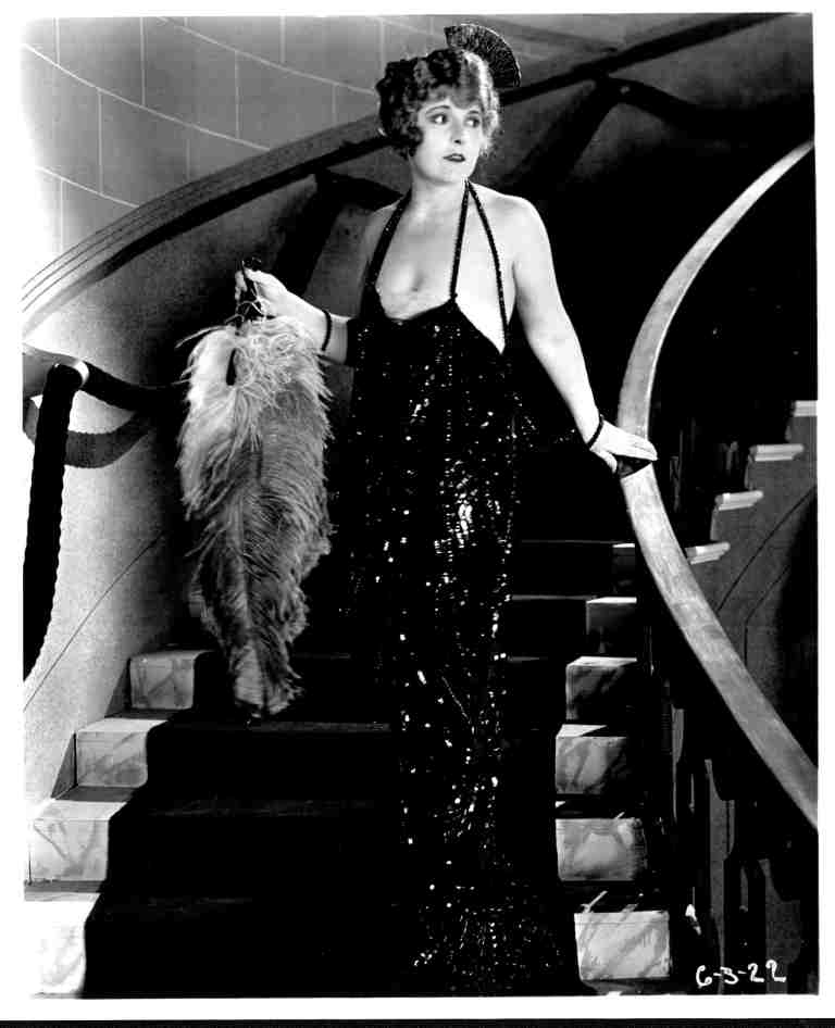 Black-and-white photo of a glamorous woman in a glittering halter-neck evening gown standing on a curved staircase, holding a feathered boa and looking off to the side.