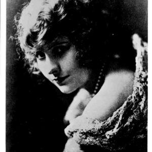 Black-and-white portrait of a woman with short curls, looking downward, wearing an off-shoulder lace dress and a pearl necklace.