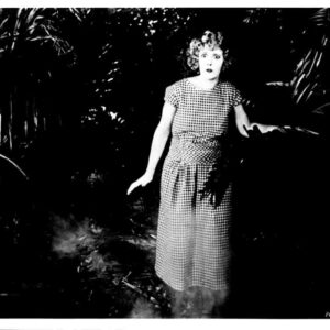 Black-and-white photo of a surprised woman in a checkered dress standing in dense foliage outside the frame.