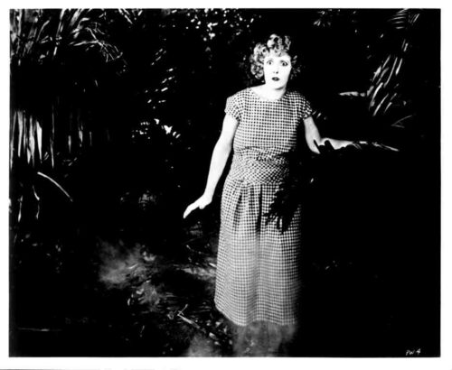 Black-and-white photo of a surprised woman in a checkered dress standing in dense foliage outside the frame.