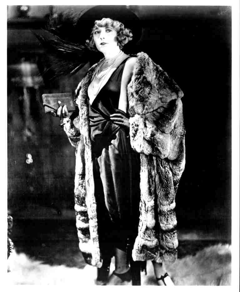 Black-and-white photo of a glamorous woman in a satin dress, pearls, fur wrap, and cloche hat, holding a clutch.
