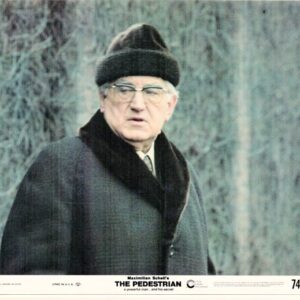 Still from the film The Pedestrian featuring Maximilian Schell, an older man in a dark overcoat, fur hat, and glasses outdoors.