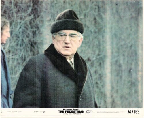 Still from the film The Pedestrian featuring Maximilian Schell, an older man in a dark overcoat, fur hat, and glasses outdoors.