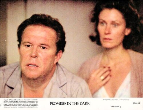 Movie still: a man in light pajamas looks tense, with a worried woman behind him in a dim room.